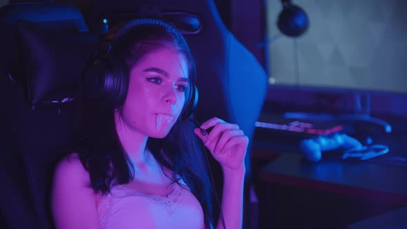 Young Woman Sitting in Neon Gaming Club and Blowing a Bubblegum - Wearing Big Headphones and Talking