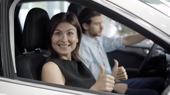 Excited Beautiful Woman Smiling at Camera and Showing Thubms Up Pointing at Man Sitting on Driver's