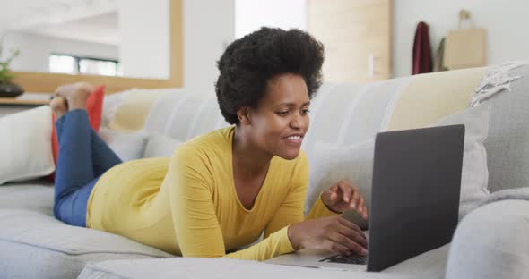 Happy african american woman laying on sofa using laptop