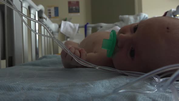 Baby in a hospital crib attached to an IV line sucking on a pacifier, starts to cry.  Shot in high s