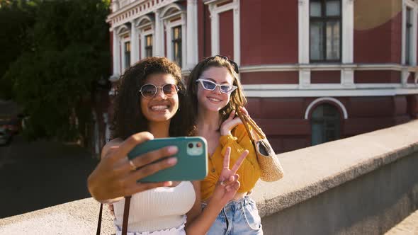 Two Young Girls in Sunglasses are Smiling Showing Victory Sign Taking Selfie on Smartphone