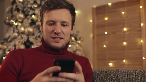 Attractive Man Using Smartphone Sitting on Couch, Messaging, Smiling in Decorated Apartment