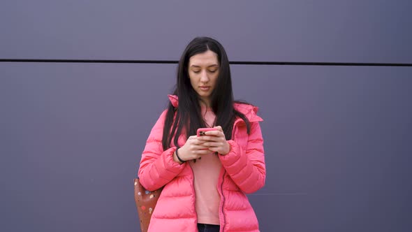 Attractive Girl with Long Brown Hair Texting on Smartphone with Her Back Against a Wall