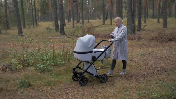 Young Mom Walking in the Park with a Stroller and Uses Phone