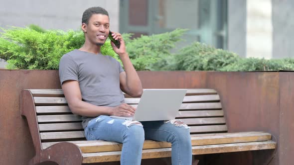 Young African Man with Laptop Talking on Smartphone on Bench 