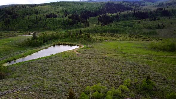 A drone shot coming to a pond with a green meadow surround it. Green pine trees and a mountain are s