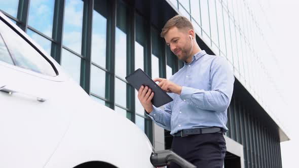 Young European Businessman with Wireless Headphones Using Tablet Near Electric Car Charging the