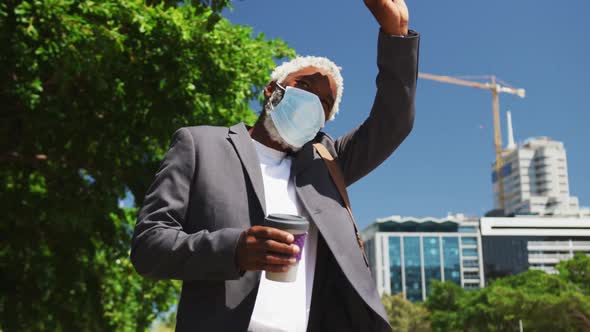 African american senior man wearing face mask holding smartphone and coffee cup hailing taxi on the