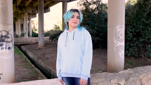 Smiling blue haired teenage girl wearing hoodie and staying against bridge pillars