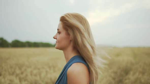 Closeup View of Attractive Young Blond Woman in a Blue Dress Walking Through Golden Wheat Field