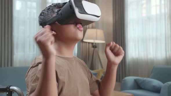 Excited Asian Boy Sitting In A Wheelchair While Wearing Virtual Reality Goggles And Dancing At Home