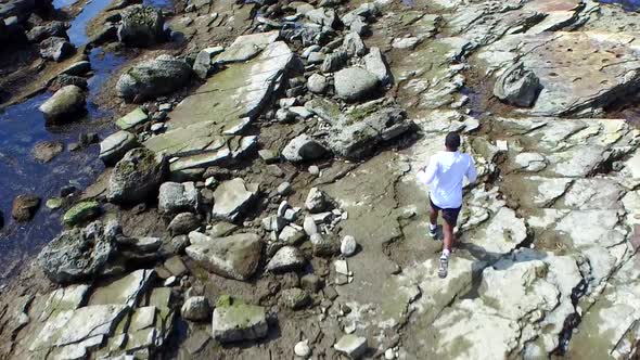 Aerial birds eye view shot of a young man running on a rocky ocean beach shoreline.