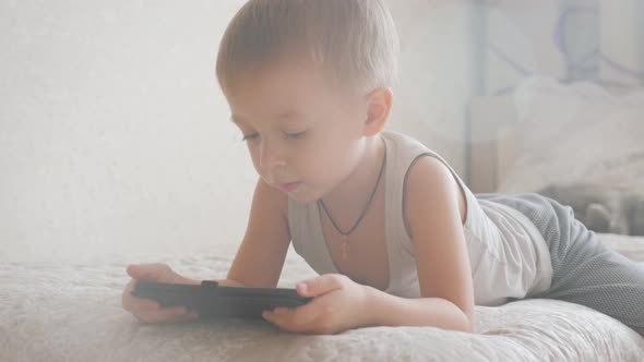 Little Boy Sitting on a Bed and Plays on the Tablet in the Room