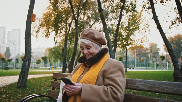 Grayhaired Woman Typing Message on Mobile Phone and Smiling While Sitting on Wooden Bench in Autumn