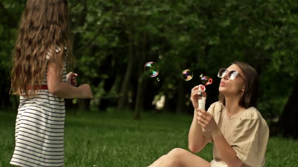 Beautiful Young Mother Together with Her Daughter in Nature Making Soap Bubbles and Laughing