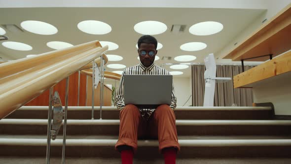 Afro-American Man Sitting on Stairs in Library and Using Laptop