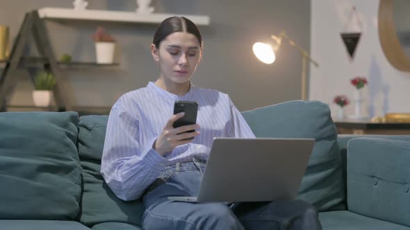 Hispanic Woman with Laptop using Smartphone on Sofa