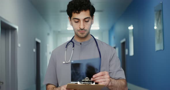 A Doctor with a Phonendoscope Walks Along the Corridor and Looks Into a Notebook
