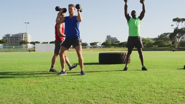 Diverse group of two fit men and male trainer exercising outdoors squatting and lifting dumbbells