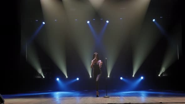 A Young Woman in a Black Suits Sings a Song and Dances on a Dark Stage.