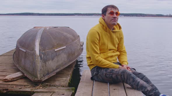 Young Man in Bright Glasses and Hoodie Sitting on Wooden Pier and Looking Around on Background of