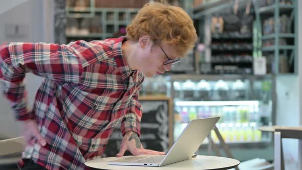 Young Redhead Man with Back Pain Using Laptop in Cafe