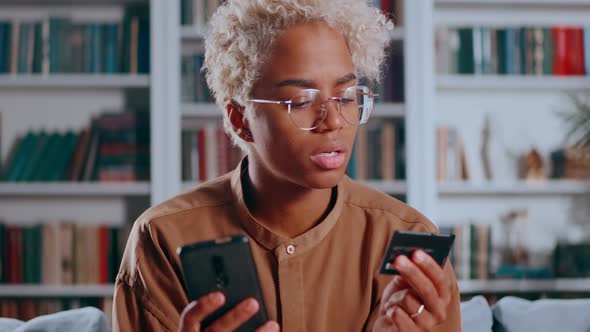 Young African American Woman Makes Online Purchases in Mobile Phone Sits on Sofa