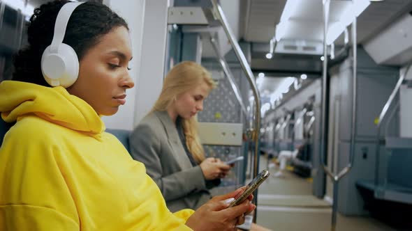 Two Young Girls are Sitting in Modern Metro Looking at Phones Listening Music