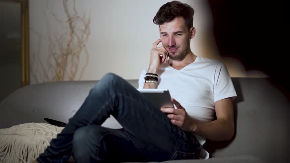 A young man with a stubble and a modern hairstyle, wearing a white t-shirt and jeans, sitting at hom