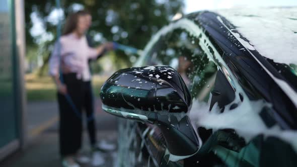 Closeup Black Car Side View Mirror with White Foam and Blurred Couple Washing Vehicle at Background