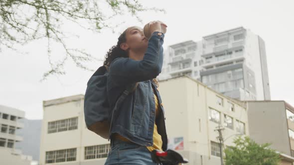 Happy biracial woman in city, wearing backpack and drinking takeaway coffee