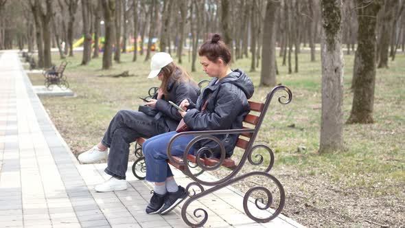 a Woman and a Girl in Black Jackets Sit on a Bench with Smartphones