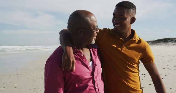 African american senior father and teenage son standing on beach embracing and talking