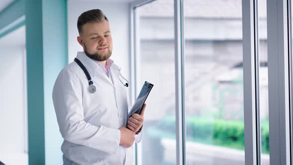 Friendly Male Doctor Standing Next Large Window at Hospital Corridor Holding Folder in His Hands