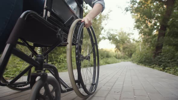 Disabled woman pushes wheelchair wheel with her hand while ride along path in park, front view.