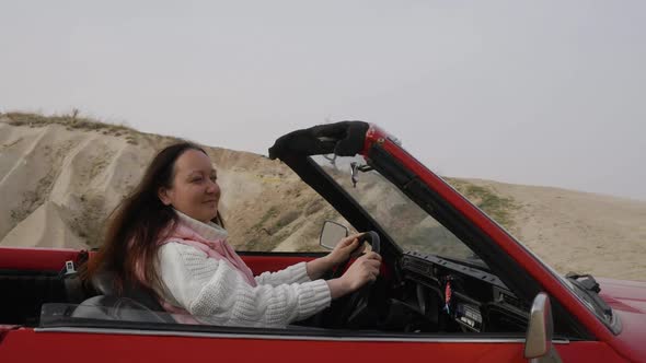 Brunette Woman Sit Behind Wheel of Red Classic Convertible Car in Desert Valley Looking Around