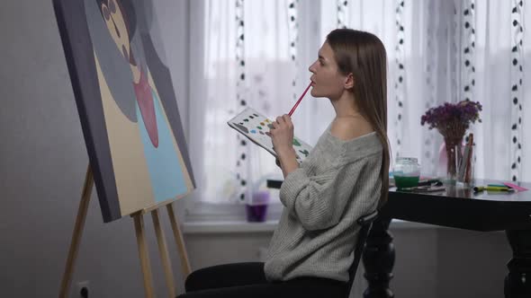 Side View Gorgeous Female Painter Making Strokes with Acrylic Paint Brush Sitting at Picture