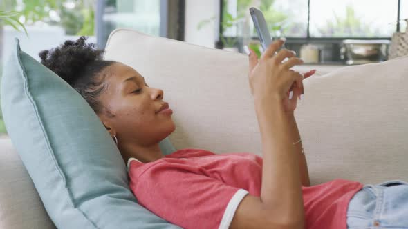 Video of happy african american woman lying on sofa and using smartphone