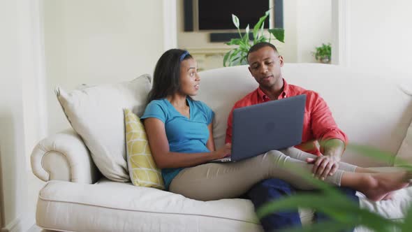 Happy biracial couple sitting on sofa with laptop and talking