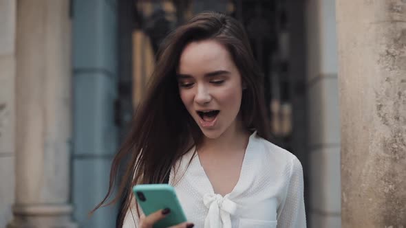 Pretty Young Brunette Girl Walking at Cosy Old Town Architecture Backgroud While Using Her Phone