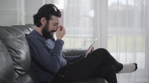 Side View of Tired Bearded Caucasian Man Rubbing Eyes, Taking on Eyeglasses and Using Tablet