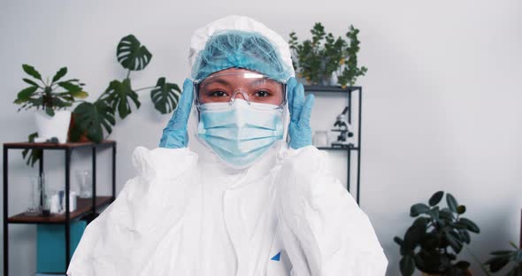 Young Multiethnic Female Doctor Wears Disposable Medical Suit and Mask Adjusting Eyeglasses During