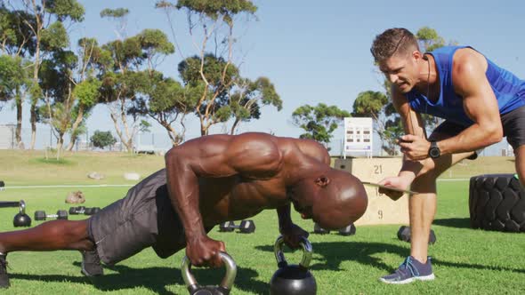 Two diverse fit men exercising outdoors, one doing push ups on kettlebells while the other times him