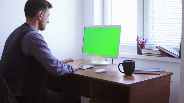 Young Man is Working on a Computer with a Mockup Green Screen at the Table By the Window