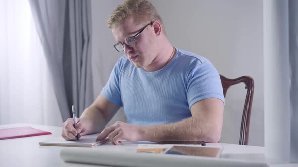 Portrait of Confident Caucasian Businessman in Eyeglasses Sitting at the Table and Taking Notes