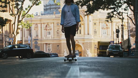 Fellow in Casual Outfit and Hat is Riding Skateboard Along City Street Surrounded By Picturesque