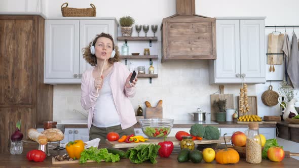 Pregnant Woman Dancing And Singing In Headphones With Mobile Phone On Kitchen.