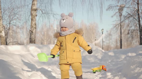 A toddler in warm yellow winter clothes runs towards the camera with a toy shovel in his hand.