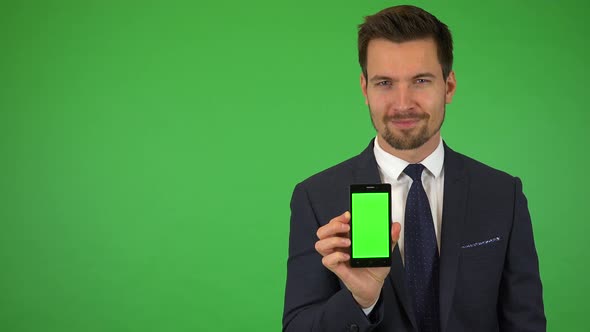 A Young Businessman Shows a Smartphone with Green Screen To the Camera and Smiles - Green Screen