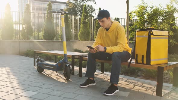 A Young Food Delivery Man Waits for the Order Sitting on a Bench with a Thermal Bag and an Electric